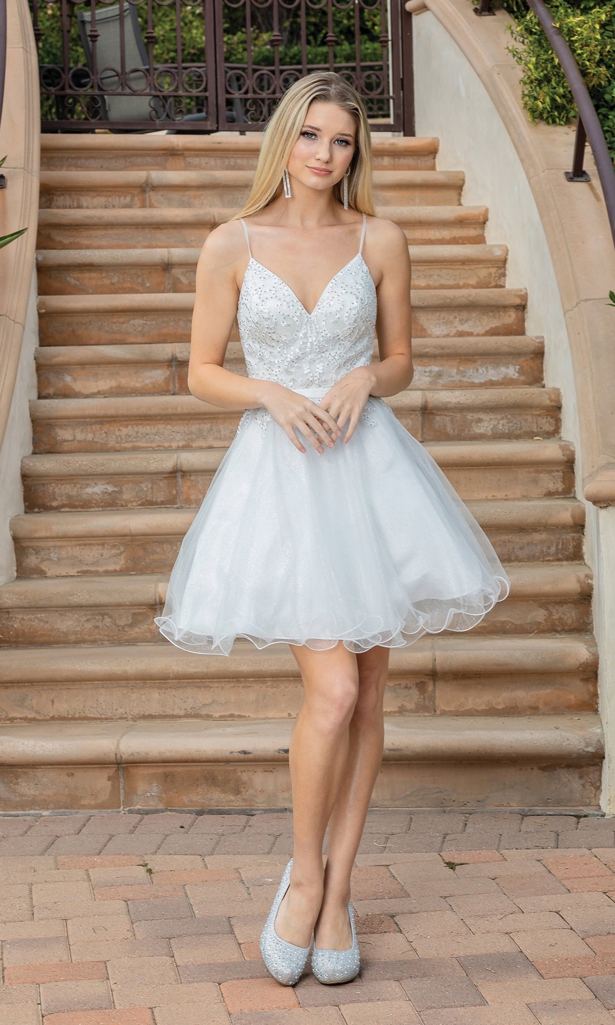 A young woman in the Lace-Up-Back Short Babydoll Homecoming Dress 3315 with a beaded corset bodice and heels poses on an outdoor staircase, hands clasped in front of her.